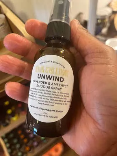 A hand holding a small lavender and amethyst unwind smudge spray